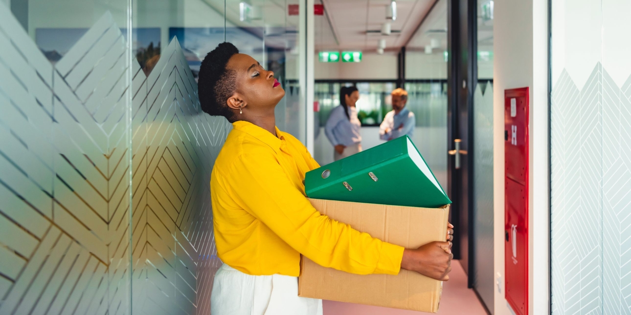 A black woman with dark hair, wearing a yellow jumper and white bottoms leans against a patterned glass panel in a corridor whilst holding a box full of files,there are other people blurred in the background